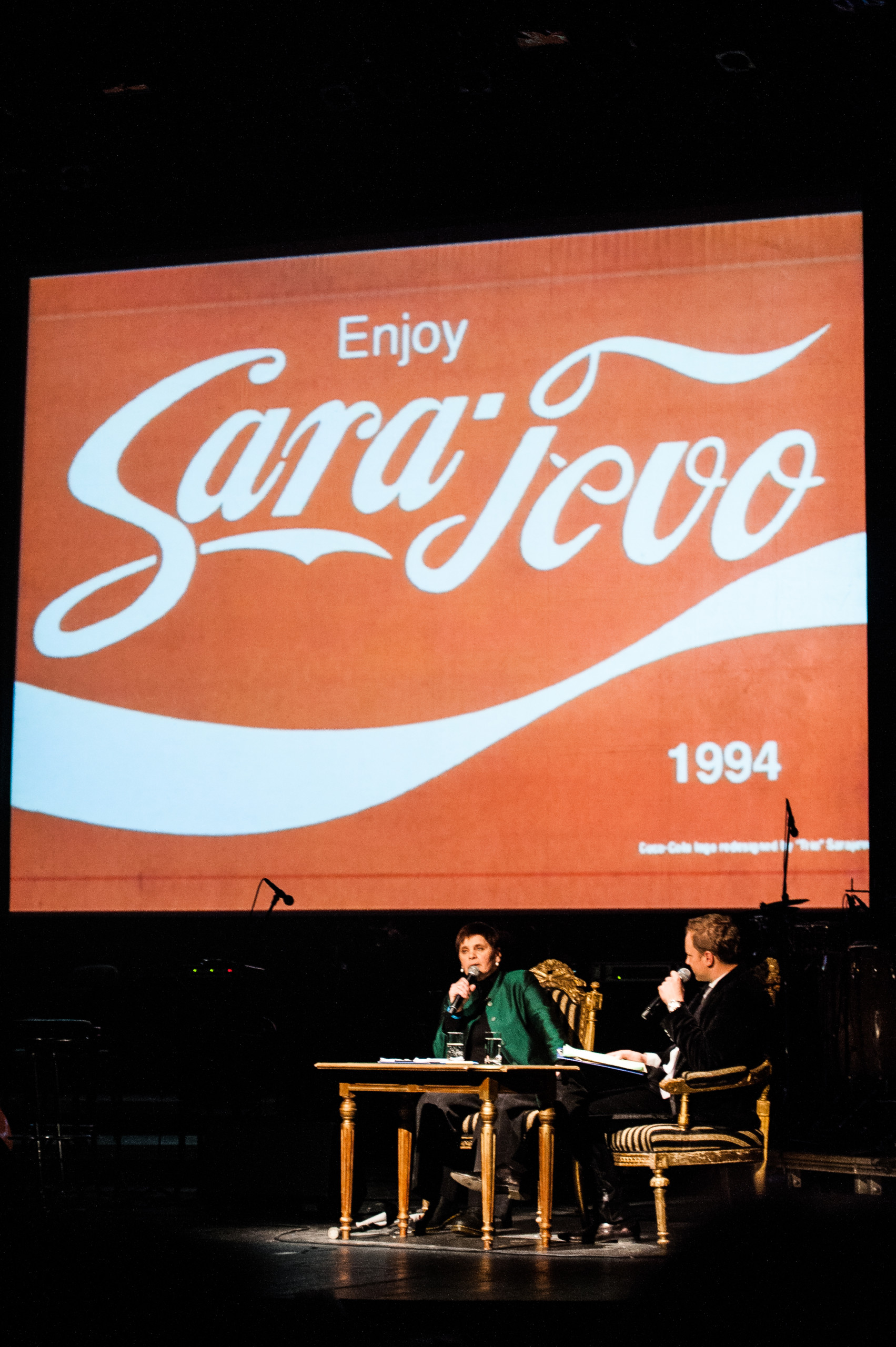 Zdjęcie Janiny z Maciejem Stuhrem a za nimi napis "Enjoy Sarajevo" stylizowany na logo Coca-Coli.